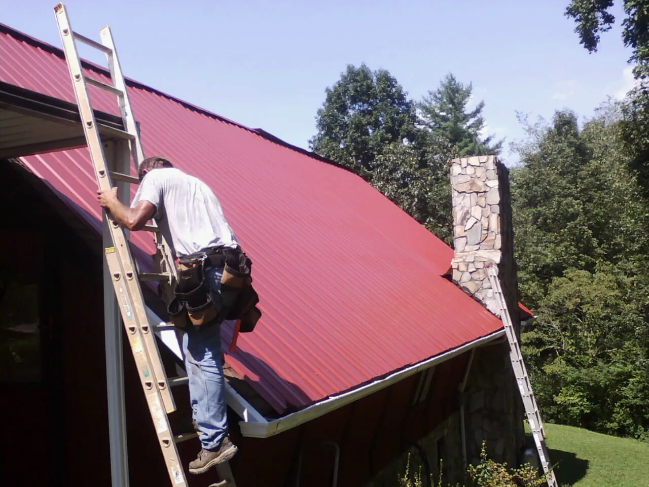 Roofer on a ladder beside a red metal roof and stone chimney — Soffit & Fascia Repair by Roofing Brothers in Yachats